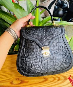 Hình ảnh túi xách HM-24 da cá sấu màu đen, thiết kế quý phái, khóa vàng nổi bật, trên bàn gỗ với lá xanh và xe máy phía sau.