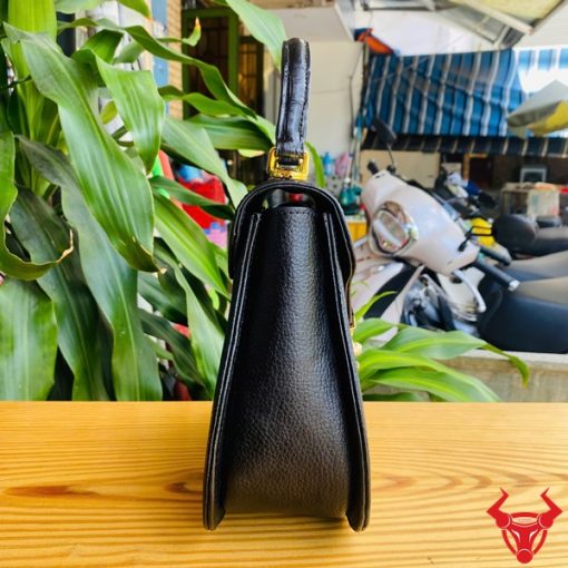 Hình ảnh túi xách da cá sấu HM-24 màu đen, kiểu dáng sang trọng, khóa vàng, đặt trên bàn gỗ với lá xanh và xe máy mờ phía sau.