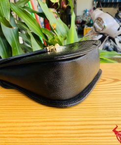 Hình ảnh túi xách HM-24 da cá sấu màu đen, thiết kế quý phái, khóa vàng lấp lánh, trên bàn gỗ với lá xanh và xe máy phía sau.