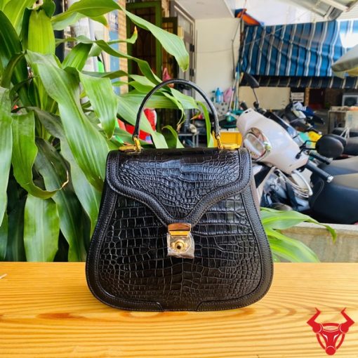 Hình ảnh túi xách da cá sấu trơn bụng HM-24 màu đen, thiết kế quý phái, khóa vàng, đặt trên bàn gỗ với nền lá xanh và xe máy mờ phía sau.