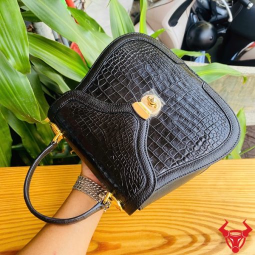 Hình ảnh túi xách da cá sấu HM-24 màu đen, kiểu dáng quý phái, khóa vàng, đặt trên bàn gỗ với lá xanh và xe máy mờ phía sau.