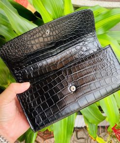 Tay cầm ví nữ ba gấp da cá sấu màu đen, nền lá xanh nổi bật.