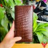 Tay nam cầm ví da bụng cá sấu nâu đất bóng đẹp giữa nền cây.