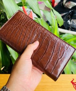 Ví da cá sấu màu nâu đất trên nền bàn gỗ, tay người cầm chắc chắn.