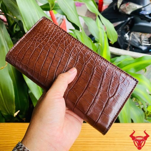 Ví da cá sấu màu nâu đất trên nền bàn gỗ, tay người cầm chắc chắn.