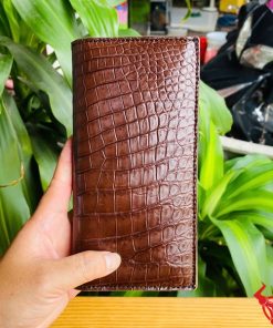 Tay nam cầm ví da bụng cá sấu nâu đất bóng đẹp giữa nền cây.