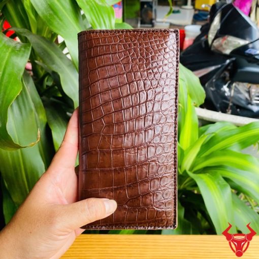 Tay nam cầm ví da bụng cá sấu nâu đất bóng đẹp giữa nền cây.