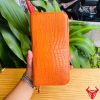 Ví cam da cá sấu cầm tay trên nền lá xanh và bàn gỗ vàng.