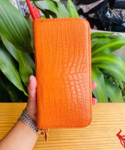 Ví cam da cá sấu cầm tay trên nền lá xanh và bàn gỗ vàng.