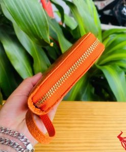 Tay người cầm ví da cá sấu cam rực rỡ, nền lá xanh hài hòa.