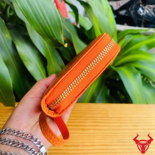 Tay người cầm ví da cá sấu cam rực rỡ, nền lá xanh hài hòa.
