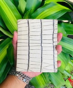 Tay nam cầm ví da cá sấu trắng viền đen nổi bật giữa nền lá xanh.