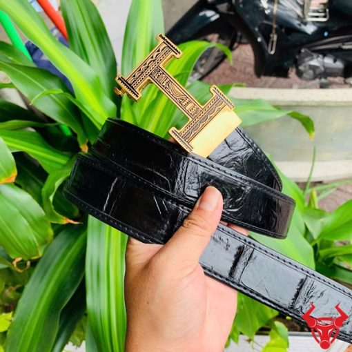 Hình ảnh thắt lưng TSK103 màu đen, da cá sấu cao cấp, đầu khóa ray thép không gỉ, cầm trên tay với cây xanh phía sau.