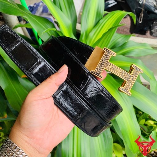 Hình ảnh thắt lưng TSK103 màu đen, da cá sấu sang trọng, đầu khóa ray thép không gỉ, cầm trên tay với nền cây xanh và mô tô.