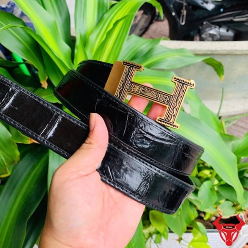 Hình ảnh thắt lưng TSK103 màu đen, da cá sấu cao cấp, đầu khóa ray thép không gỉ, cầm trên tay với cây xanh và mô tô phía sau.