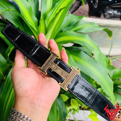 Hình ảnh thắt lưng da cá sấu TSK103 màu đen, đầu khóa ray thép không gỉ, được cầm trên tay trước nền cây xanh rực rỡ.