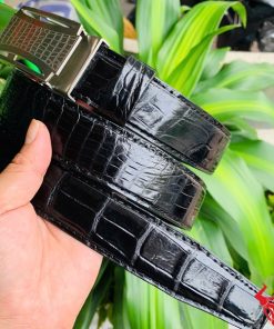 Hình ảnh thắt lưng TSK32 màu đen, da cá sấu cao cấp, mặt khóa lăn inox, cầm trên tay với cây xanh phía sau.