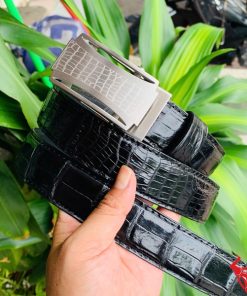 Hình ảnh thắt lưng da cá sấu TSK32 màu đen, mặt khóa lăn inox cao cấp, được cầm trên tay trước nền cây xanh và mô tô.