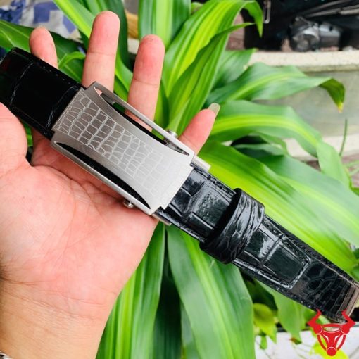 Hình ảnh thắt lưng TSK32 màu đen, da cá sấu cao cấp, mặt khóa lăn inox, cầm trên tay trước nền cây xanh.