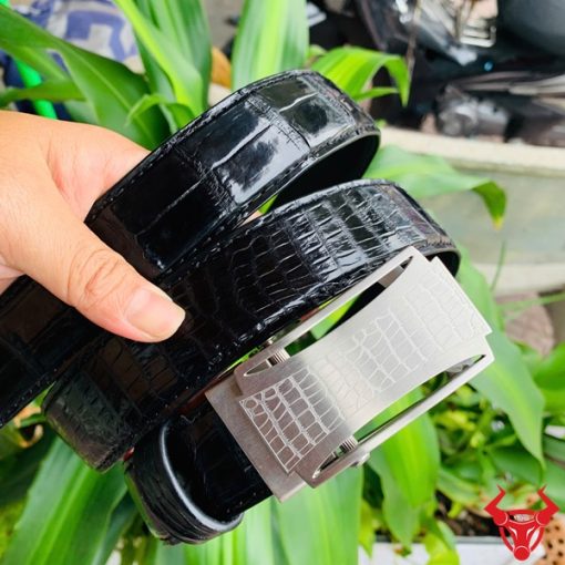 Hình ảnh thắt lưng TSK32 màu đen, da cá sấu cao cấp, mặt khóa lăn inox, cầm trên tay với cây xanh và mô tô phía sau.