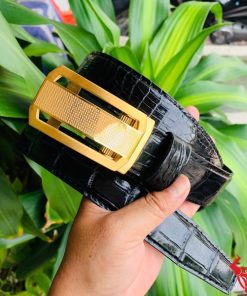 Hình ảnh thắt lưng TSK33 màu đen, da cá sấu cao cấp, khóa tỳ thép vàng, cầm trên tay với cây xanh phía sau.
