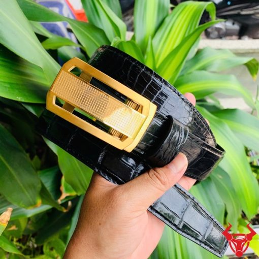 Hình ảnh thắt lưng TSK33 màu đen, da cá sấu cao cấp, khóa tỳ thép vàng, cầm trên tay với cây xanh phía sau.