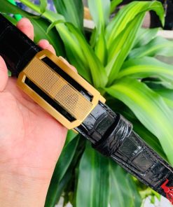 Hình ảnh thắt lưng da cá sấu TSK33 màu đen, khóa tỳ thép màu vàng, được cầm trên tay trước nền cây xanh rực rỡ.