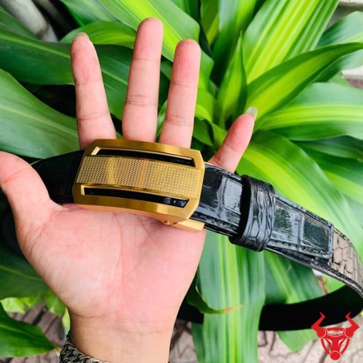 Hình ảnh thắt lưng TSK33 màu đen, da cá sấu cao cấp, khóa tỳ thép vàng, cầm trên tay với cây xanh và mô tô phía sau.