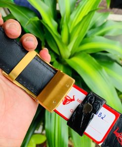 Hình ảnh thắt lưng TSK33 màu đen, da cá sấu sang trọng, khóa tỳ thép vàng, cầm trên tay với cây xanh phía sau.