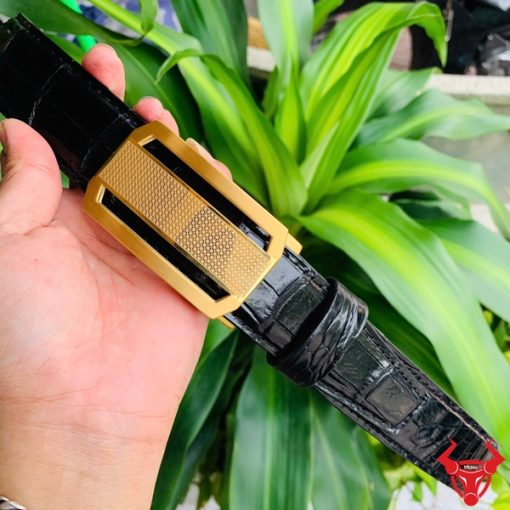 Hình ảnh thắt lưng da cá sấu TSK33 màu đen, khóa tỳ thép màu vàng, được cầm trên tay trước nền cây xanh rực rỡ.