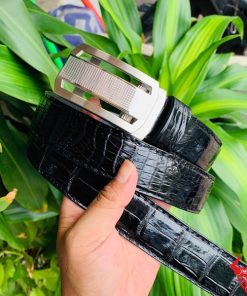 Hình ảnh thắt lưng TSK33 màu đen, da cá sấu cao cấp, khóa tỳ thép trắng, cầm trên tay với cây xanh phía sau.