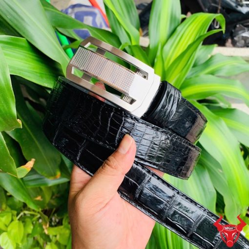 Hình ảnh thắt lưng TSK33 màu đen, da cá sấu cao cấp, khóa tỳ thép trắng, cầm trên tay với cây xanh phía sau.