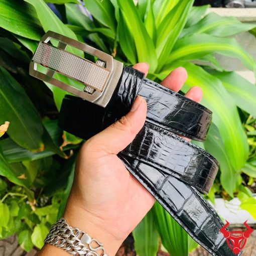 Hình ảnh thắt lưng TSK33 màu đen, da cá sấu sang trọng, khóa tỳ thép trắng, cầm trên tay với nền cây xanh và mô tô.