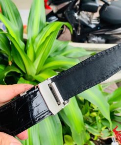 Hình ảnh thắt lưng TSK33 màu đen, da cá sấu cao cấp, khóa tỳ thép trắng, cầm trên tay trước nền cây xanh.