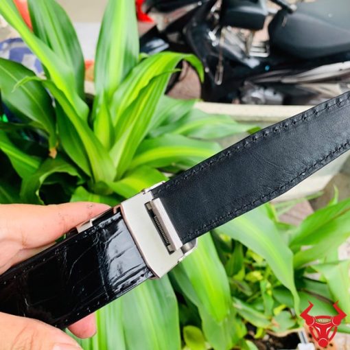 Hình ảnh thắt lưng TSK33 màu đen, da cá sấu cao cấp, khóa tỳ thép trắng, cầm trên tay trước nền cây xanh.
