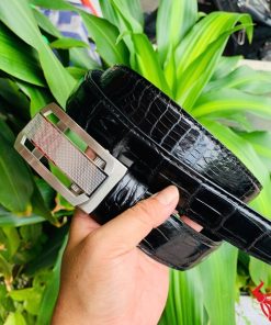 Hình ảnh thắt lưng TSK33 màu đen, da cá sấu cao cấp, khóa tỳ thép trắng, cầm trên tay với cây xanh và mô tô phía sau.