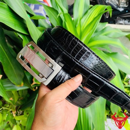 Hình ảnh thắt lưng TSK33 màu đen, da cá sấu cao cấp, khóa tỳ thép trắng, cầm trên tay với cây xanh và mô tô phía sau.