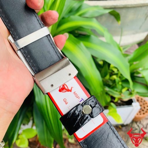 Hình ảnh thắt lưng TSK33 màu đen, da cá sấu sang trọng, khóa tỳ thép trắng, cầm trên tay với cây xanh phía sau.