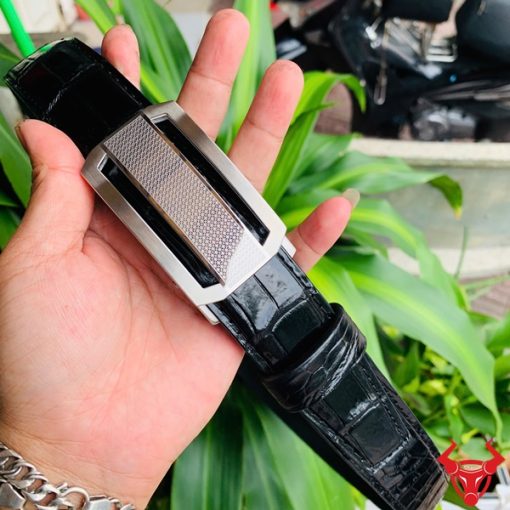 Hình ảnh thắt lưng da cá sấu TSK33 màu đen, khóa tỳ thép trắng cao cấp, được cầm trên tay trước nền cây xanh rực rỡ.