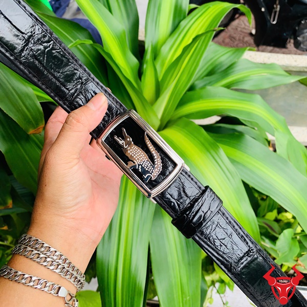 Sang trọng với Thắt Lưng Da Cá Sấu TSK73 Hình ảnh thắt lưng TSK73 màu đen, da cá sấu cao cấp, mặt khóa ray trượt inox, cầm trên tay với cây xanh phía sau.