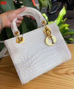 Hình ảnh túi xách nữ da bụng cá sấu TDA8 màu bạch tạng, thiết kế quý phái, khóa vàng lấp lánh, đặt trên bàn gỗ với cây xanh và mô tô.