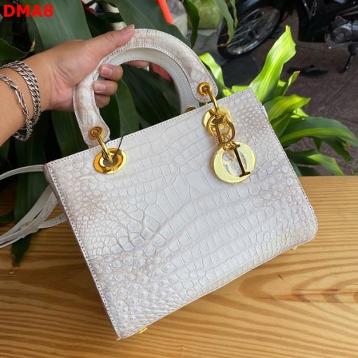 Hình ảnh túi xách nữ da bụng cá sấu TDA8 màu bạch tạng, thiết kế quý phái, khóa vàng lấp lánh, đặt trên bàn gỗ với cây xanh và mô tô.