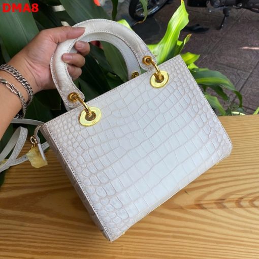 Hình ảnh túi xách TDA8 màu bạch tạng, da bụng cá sấu sang trọng, khóa vàng lấp lánh, đặt trên bàn gỗ với cây xanh và mô tô.