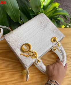 Hình ảnh túi xách TDA8 màu bạch tạng, da bụng cá sấu cao cấp, khóa vàng lộng lẫy, đặt trên bàn gỗ với nền cây xanh.