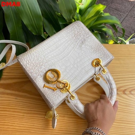 Hình ảnh túi xách TDA8 màu bạch tạng, da bụng cá sấu cao cấp, khóa vàng lộng lẫy, đặt trên bàn gỗ với nền cây xanh.
