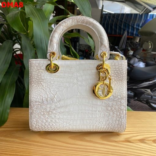 Hình ảnh túi xách TDA8 màu bạch tạng, da bụng cá sấu cao cấp, khóa vàng nổi bật, đặt trên bàn gỗ với cây xanh phía sau.