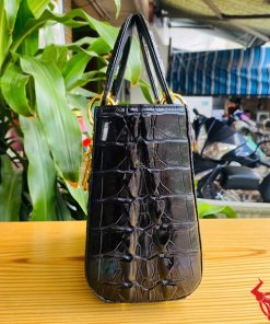 Hình ảnh túi xách TDA4 màu đen, da cá sấu gù cao cấp, khóa vàng nổi bật, đặt trên bàn gỗ với cây xanh phía sau.