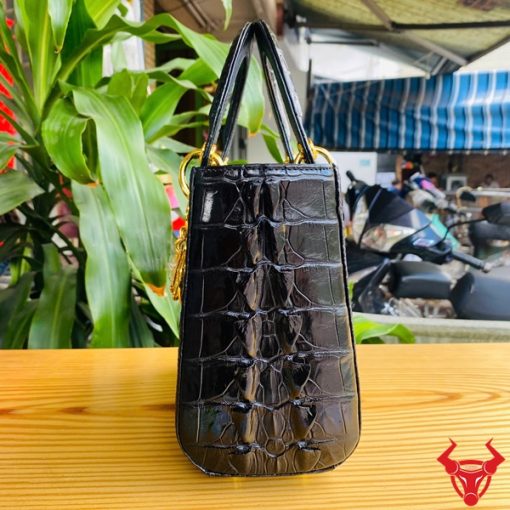 Hình ảnh túi xách TDA4 màu đen, da cá sấu gù cao cấp, khóa vàng nổi bật, đặt trên bàn gỗ với cây xanh phía sau.