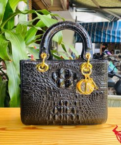 Hình ảnh túi xách nữ da cá sấu gù TDA4 màu đen, thiết kế cao cấp, khóa vàng lấp lánh, đặt trên bàn gỗ với cây xanh và mô tô.