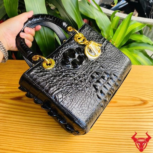 Hình ảnh túi xách TDA4 màu đen, da cá sấu gù cao cấp, khóa vàng lộng lẫy, đặt trên bàn gỗ với nền cây xanh.